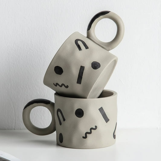 Two gray ceramic mugs with black abstract face designs on a white background