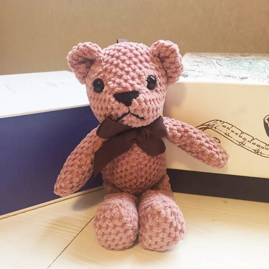 Pink knitted teddy bear with a brown bow tie on a wooden surface.
